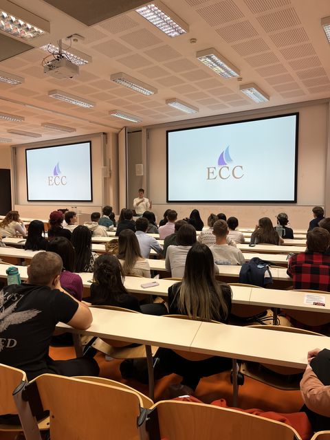 A packed lecture hall facing a speaker, with the ECC logo on two big screens